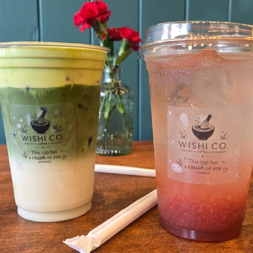 Two Wishli Co. iced drinks, including a layered matcha latte and pink fruit beverage, sit on a wooden table with flowers.