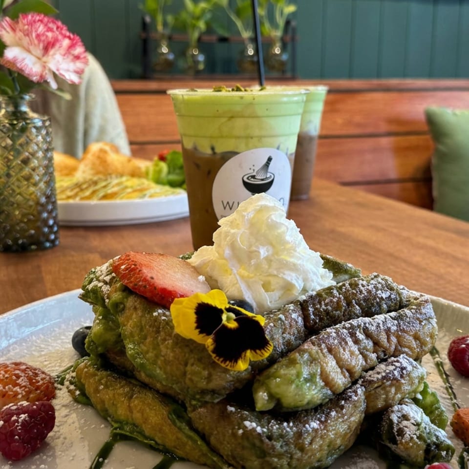 Stack of green waffles dusted with powdered sugar, topped with whipped cream, strawberry, edible flower, and berries on a café table.