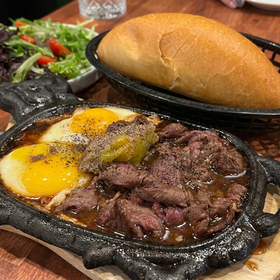 Sizzling beef steak with two sunny-side-up eggs and peppers in a cast-iron skillet, served with bread and salad.