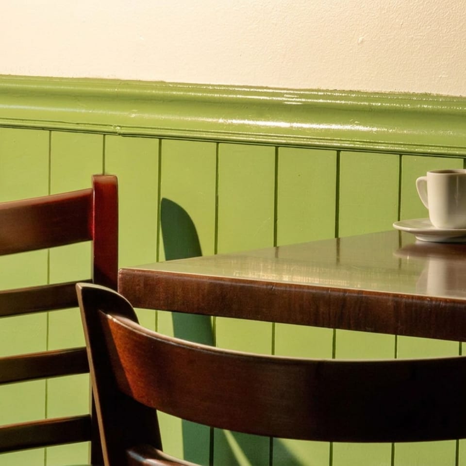 Cozy restaurant table with two coffee cups, wooden chairs, and green paneled walls illuminated by warm sunlight.
