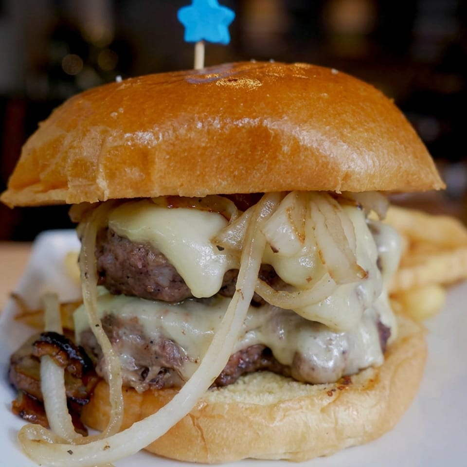 Double cheeseburger with grilled onions on a toasted bun, served with fries on a white plate in a restaurant.