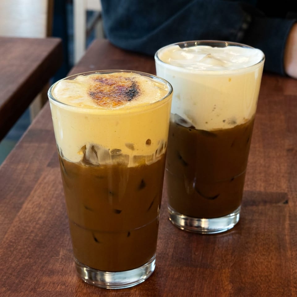 Two iced coffee drinks topped with frothy cream sit on a wooden restaurant table beside a water glass.